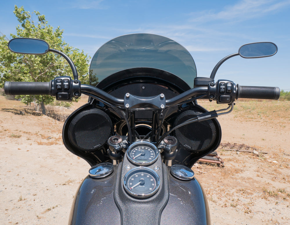 A close-up, eye-level view of the handlebars, fairing, and gauges of a dark gray RWD V-Twin motorcycle, parked outdoors on a dirt path with a tree and blue sky in the background. The motorcycle features black handlebars with mirrors, a dark tinted windshield, and two round speakers integrated into the fairing. Two chrome-rimmed gauges are visible on the tank.