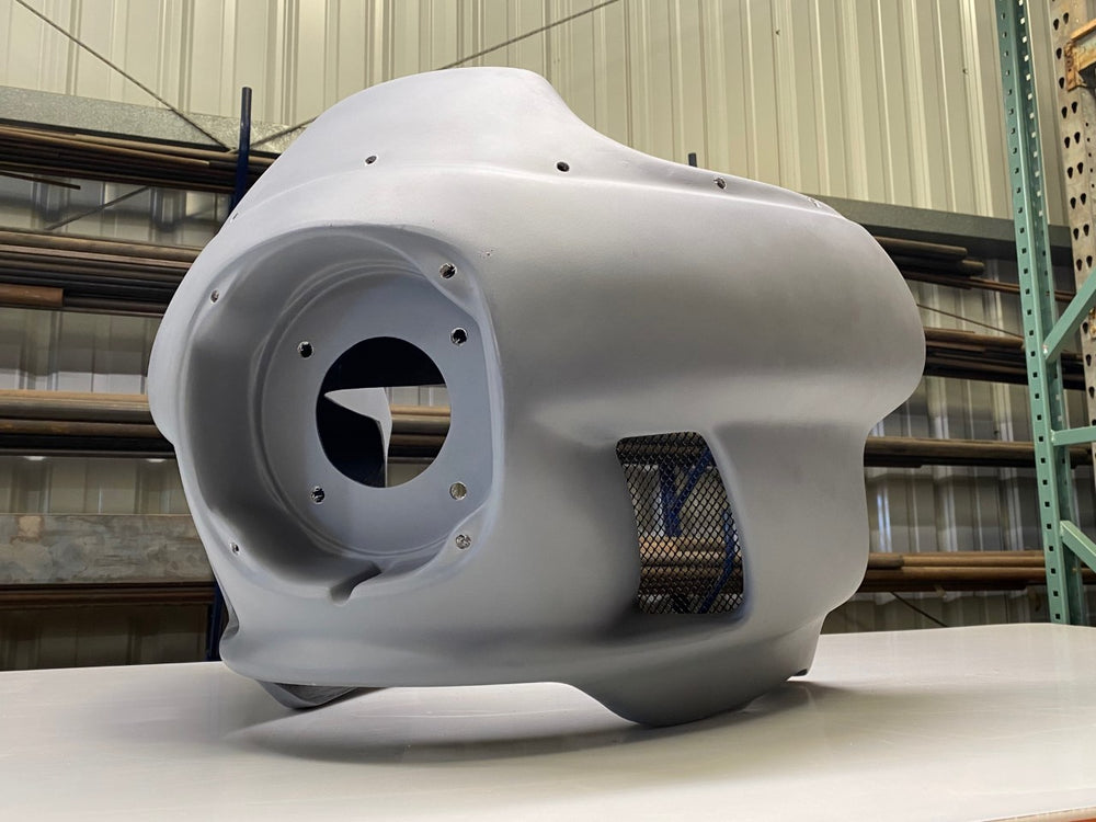 A light gray RWD V-Twin motorcycle fairing, featuring a large circular opening for a headlight and a mesh-covered vent on the side, sits on a white surface in a warehouse setting.